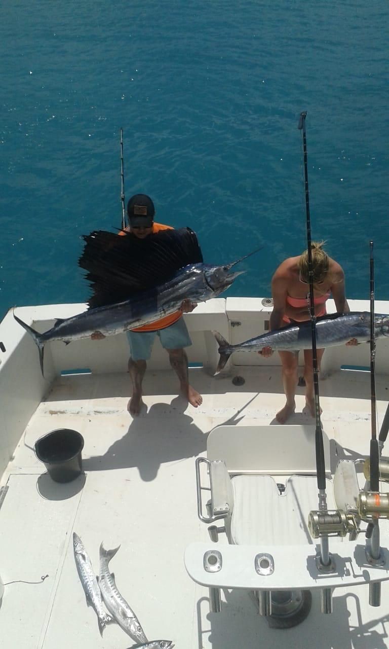 Two people on a boat holding large fish with fishing rods and ocean in the background.