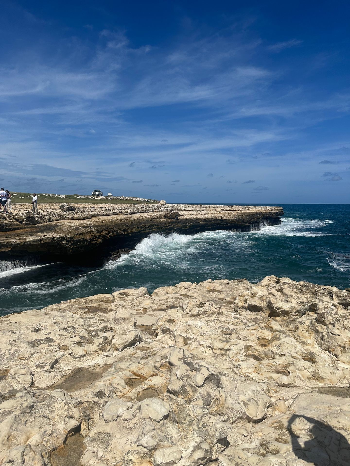 Rocky coastline with waves crashing against the shore under a blue sky with scattered clouds.