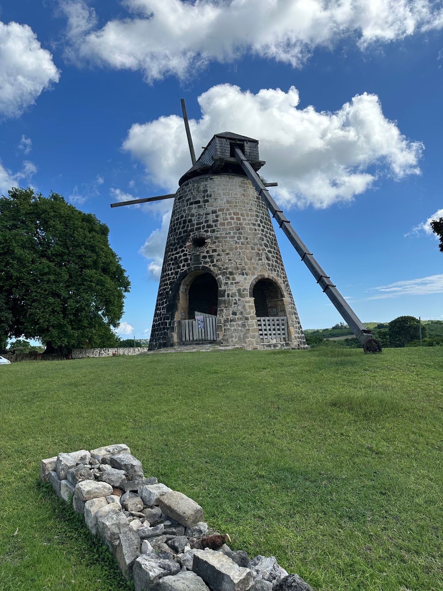 Old stone windmill with wooden blades on a grassy field under a blue sky with clouds.