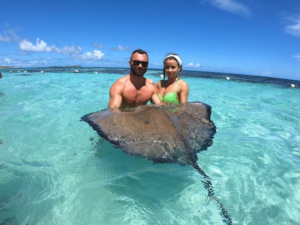 Two people holding a large stingray in clear turquoise water under a blue sky.