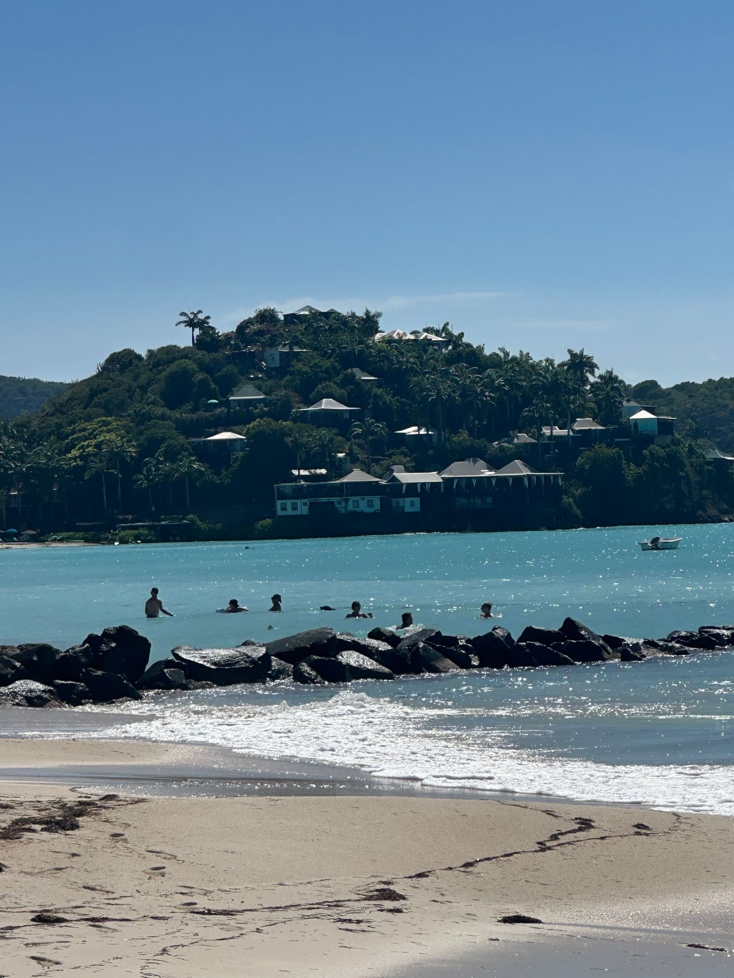 Beachfront with swimmers in the sea, rocky breakwater, and lush hillside resort in the background.