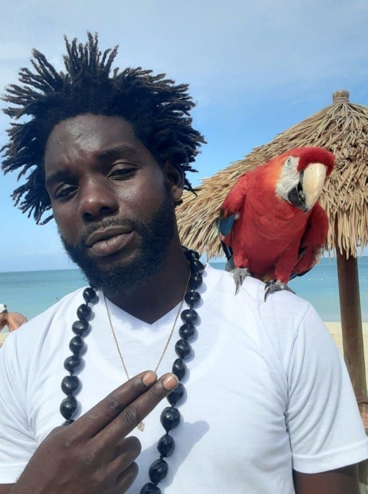 Person wearing necklace with parrot on shoulder, standing by beach and thatched umbrella.